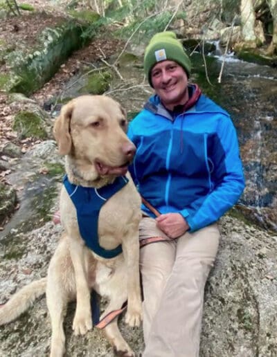 Man sitting on a rock with stream behind him and big tan dog next to him.