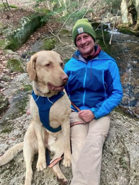 Man sitting on a rock with stream behind him and big tan dog next to him.