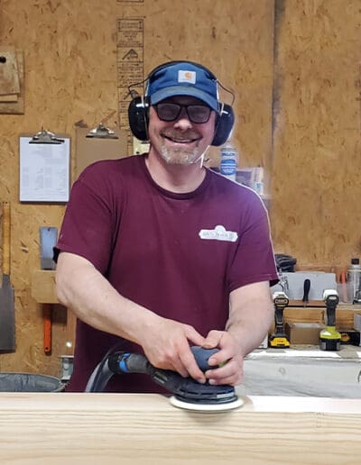 Man smiling at camera while working on sanding a timber.